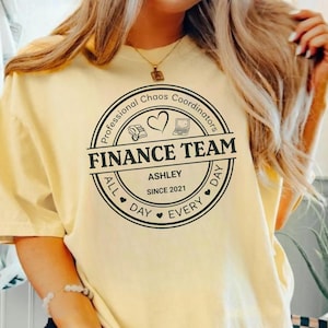 Può includere: Una t-shirt a maniche corte giallo pallido con una grafica circolare nera sul petto. Il testo recita Professional Chaos Coordinators, FINANCE TEAM, ASHLEY, SINCE 2021 e ALL DAY EVERY DAY, accompagnato da icone di una calcolatrice e un cuore.