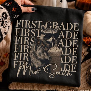 Op de afbeelding: Een zwart t-shirt met een afbeelding van een wasbeer die een zonnebril en een bandana met patroon draagt en een koffiebeker vasthoudt. De tekst FIRST GRADE is in de achtergrond herhaald in lichtbeige, met de naam Mrs. Smith eronder geschreven.