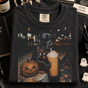 May include: A black t-shirt featuring a graphic of a black dog wearing sunglasses and a pumpkin-patterned bandana, holding an iced coffee next to a jack-o'-lantern. The shirt has a Comfort Colors label showing size L-G.