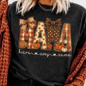 Puede incluir: Una camiseta negra con la palabra MAMA en letras grandes y estampadas con diseños de cuadros, lunares y hojas de otoño. Dos lazos marrones decoran las letras, y los nombres Kiara, Zory y Zaven aparecen escritos en cursiva debajo.