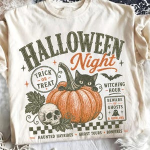May include: A cream-colored t-shirt featuring a graphic print with the text HALLOWEEN Night, Trick or Treat, Witching Hour, Beware of Ghosts & Goblins, Haunted Hayrides, Ghost Tours, and Bonfires, alongside a pumpkin, black cat, and skull.