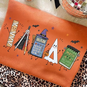 May include: An orange sweatshirt featuring a Halloween-themed graphic of school supplies as characters, including a mummy ruler, a vampire compass, a witch calculator, a ghost triangle, and a Frankenstein book with the text MATH.
