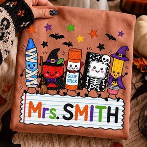 May include: A peach-colored t-shirt featuring a graphic of five cartoon school supplies dressed in Halloween costumes, including a mummy crayon, a witch apple, a vampire glue stick, a skeleton notebook, and a pencil with a witch hat, with the text Mrs. SMITH.