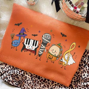 May include: An orange sweatshirt featuring a graphic print of five musical characters dressed for Halloween: a blue music note witch, a piano vampire, a mummy microphone, a pumpkin tambourine, and a ghost treble clef with small bats and stars.