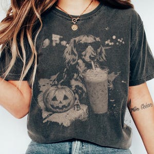 May include: A charcoal gray short-sleeve t-shirt featuring a graphic print of a long-haired dachshund wearing sunglasses, holding a plastic cup with a straw, and sitting next to a carved jack-o'-lantern pumpkin with a faded, vintage-style finish.