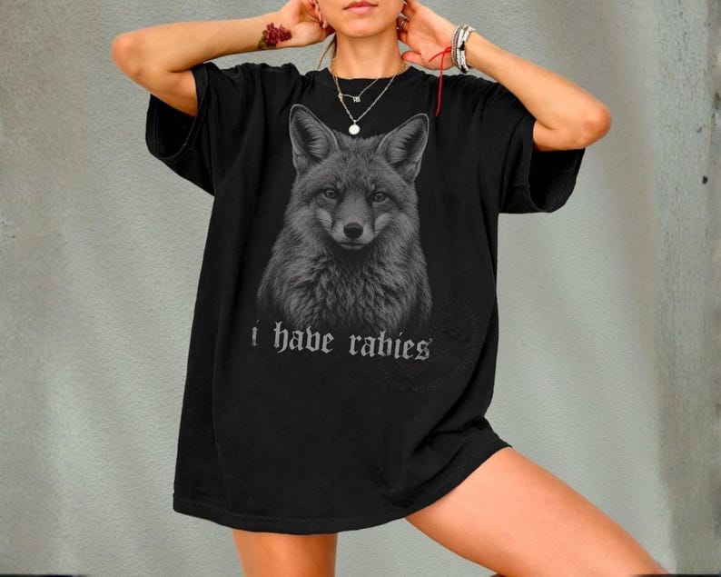 May include: A black short-sleeve t-shirt featuring a detailed grayscale illustration of a fox head centered on the chest. Below the fox graphic, the text "i have rabies" is printed in a gothic-style font in a light gray color.