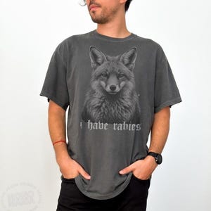 May include: A charcoal gray short-sleeve t-shirt featuring a detailed monochromatic illustration of a fox head centered on the chest. Below the fox graphic, the text "I have rabies" is printed in a stylized, gothic-inspired white font.