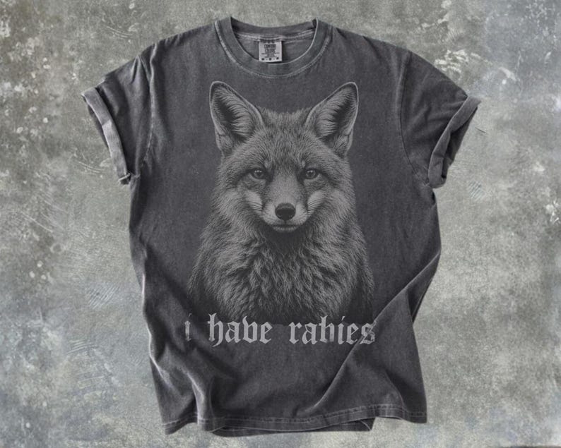 May include: A charcoal gray short-sleeve t-shirt featuring a detailed black and white illustration of a fox head centered on the chest. Below the fox graphic, the text "i have rabies" is printed in a gothic-style font. The shirt has a crew neck.