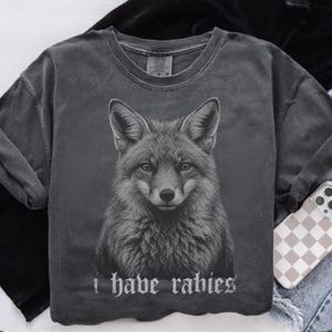 May include: A charcoal gray short-sleeve t-shirt featuring a detailed black and white illustration of a fox head centered on the chest. Below the fox, the text "i have rabies" is printed in a gothic-style font. The shirt has a crew neckline.