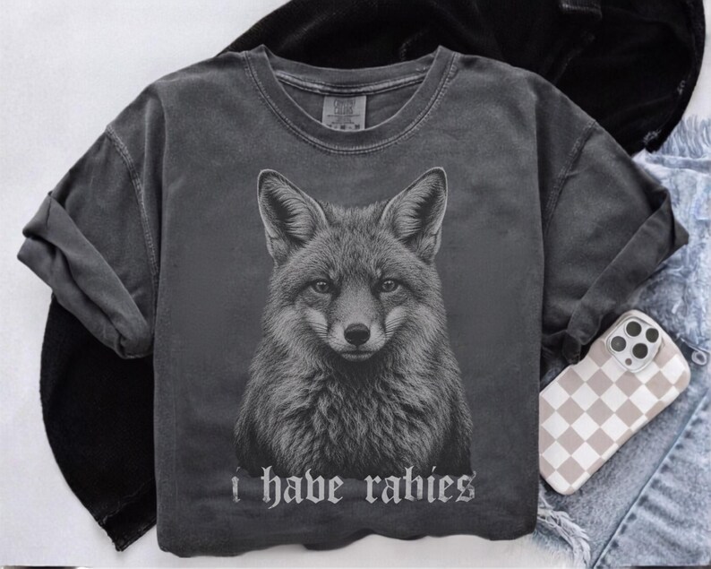 May include: A charcoal gray short-sleeve t-shirt featuring a detailed black and white illustration of a fox head centered on the chest. Below the fox, the text "i have rabies" is printed in a gothic-style font. The shirt has a crew neckline.
