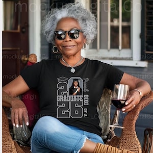 May include: A black t-shirt featuring a graduation design with the text "2026 Graduate" and a photo of a graduate. The words "Proud of A Gigi" are printed vertically on the right side. The person in the image is wearing sunglasses and holding a glass of red wine.