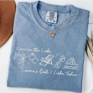 Könnte beinhalten: Ein blaues Comfort Colors T-Shirt mit dem Text "Love on the Lake" und Illustrationen eines Weinglases, Ringen, eines Strandstuhls, einer Sonnenbrille und eines Segelbootes. Eine goldene Halskette mit Anhänger liegt auf dem Shirt.