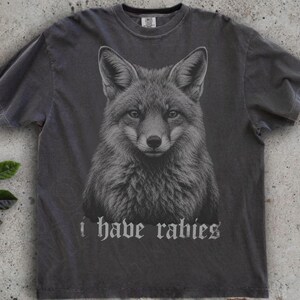 May include: A charcoal gray short-sleeve t-shirt featuring a detailed black and white illustration of a fox head centered on the chest. Below the fox graphic, the text "i have rabies" is printed in a gothic-style font in a light gray color.