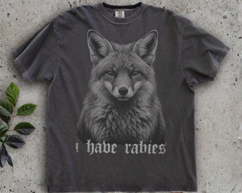 May include: A charcoal gray short-sleeve t-shirt featuring a detailed black and white illustration of a fox head centered on the chest. Below the fox graphic, the text "i have rabies" is printed in a gothic-style font in a light gray color.