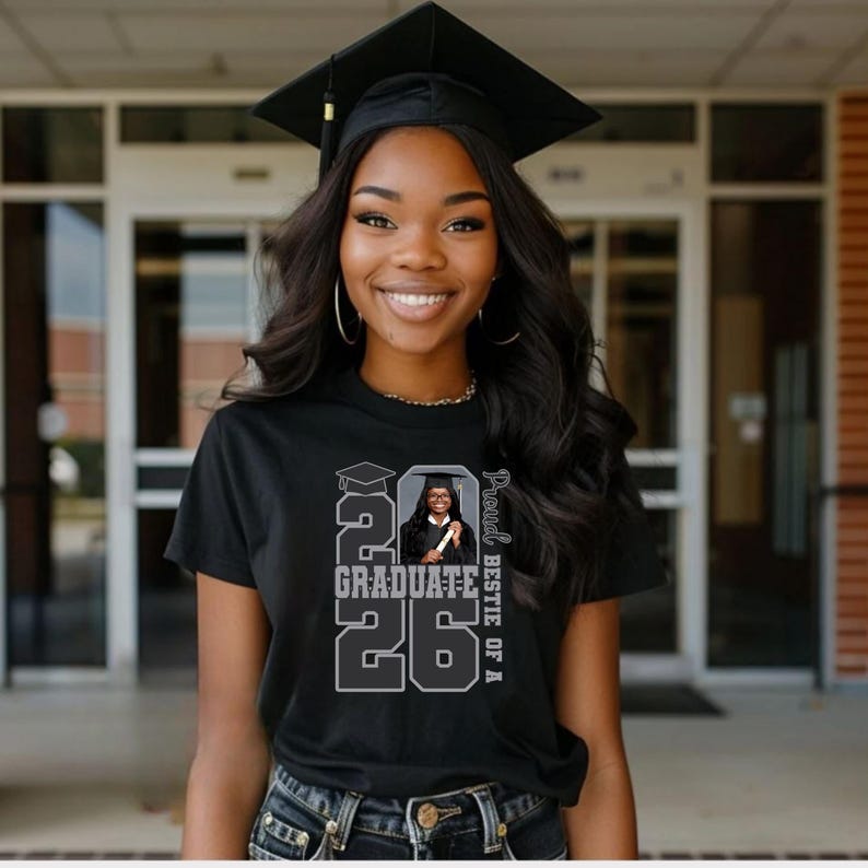 May include: A smiling person wearing a black graduation cap and a black t-shirt. The t-shirt features a graphic with the text "2026 Graduate" and a photo of a graduate. The words "Proud Bestie of A" are also printed on the shirt.