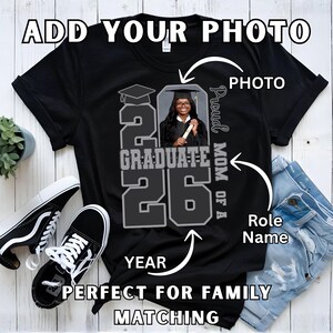 May include: Black t-shirt with a graduation design. The design includes the text "20 Graduate 26", a photo, and the words "Proud Mom of a". The shirt is next to a pair of black and white sneakers and a pair of blue jeans.