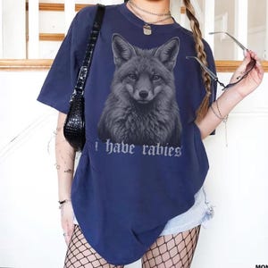 May include: A navy blue short-sleeved t-shirt featuring a detailed graphic of a fox head in the center. Below the fox, the text "i have rabies" is printed in a gothic-style font. The shirt has a crew neckline and a loose, relaxed fit.