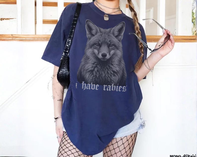 May include: A navy blue short-sleeved t-shirt featuring a detailed graphic of a fox head in the center. Below the fox, the text "i have rabies" is printed in a gothic-style font. The shirt has a crew neckline and a loose, relaxed fit.