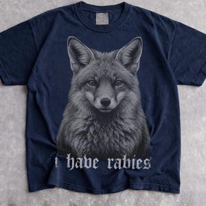 May include: A dark navy blue short-sleeve t-shirt featuring a detailed, centered graphic of a fox head in shades of gray. Below the fox illustration, the text "I have rabies" is printed in a white, gothic-style font across the chest area.