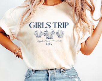 May include: Cream-colored t-shirt with the text "GIRLS TRIP" in navy blue above three seashell illustrations. Below, it reads "Myrtle Beach, SC - 2026" and "KIRA". The shirt is paired with blue jeans.