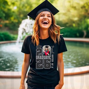 May include: A black t-shirt with the text "2026 Graduate" and a photo of a graduate in a cap and gown. The words "Proud Sister of A" are printed vertically on the right side. The person is wearing a graduation cap.