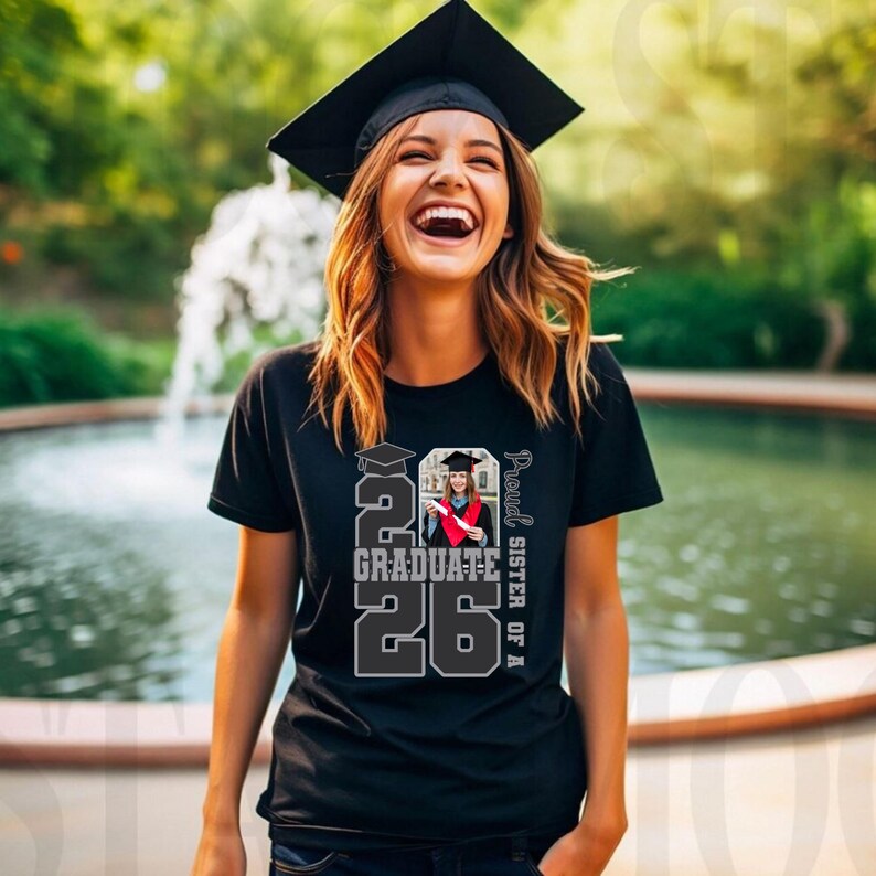 May include: A black t-shirt with the text "2026 Graduate" and a photo of a graduate in a cap and gown. The words "Proud Sister of A" are printed vertically on the right side. The person is wearing a graduation cap.