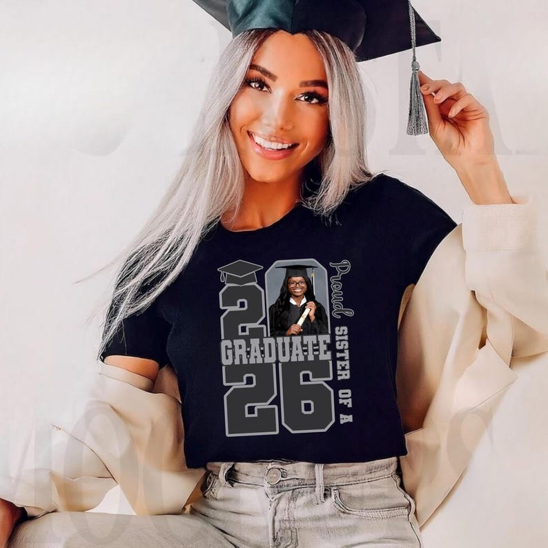 May include: Black t-shirt with a graduation theme. The shirt features the text "2026 Graduate" in large, gray block letters, with a photo of a graduate in a cap and gown. The words "Proud Sister of A" are printed vertically on the right side.