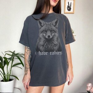 May include: A charcoal gray short-sleeve t-shirt featuring a detailed black and white illustration of a fox head centered on the chest. Below the fox, the text "i have rabies" is printed in a gothic-style font. The shirt has a crew neckline.