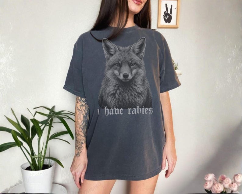 May include: A charcoal gray short-sleeve t-shirt featuring a detailed black and white illustration of a fox head centered on the chest. Below the fox, the text "i have rabies" is printed in a gothic-style font. The shirt has a crew neckline.