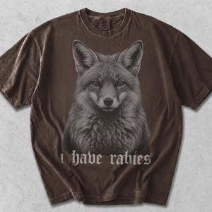 May include: A dark brown short-sleeve t-shirt featuring a detailed, centered graphic illustration of a fox head. Below the fox, the text "i have rabies" is printed in a gothic-style font. The fabric has a washed, vintage-inspired appearance.