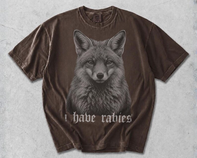 May include: A dark brown short-sleeve t-shirt featuring a detailed, centered graphic illustration of a fox head. Below the fox, the text "i have rabies" is printed in a gothic-style font. The fabric has a washed, vintage-inspired appearance.