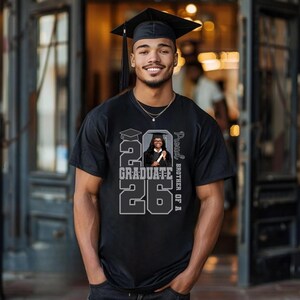 May include: A black t-shirt with a graduation design. The shirt features the text "2026 Graduate" in a bold, gray font, along with a photo of a graduate. The words "Proud Brother of A" are printed vertically on the right side of the shirt.