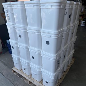 Op de afbeelding: Een grote stapel witte vierkante kunststof emmers op een houten pallet. Elke emmer is voorzien van een metalen draadhengel en een klein rond label met tekst. De emmers zijn in meerdere kolommen gestapeld, wat een uniform raster vormt.