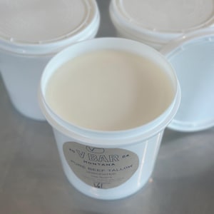 May include: A white plastic container with a label that reads "VBAR MONTANA 20 PURE BEEF TALLOW 24 UNSCENTED". The container is filled with a white substance.