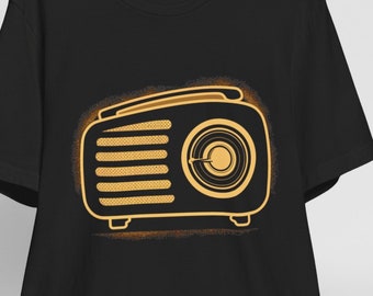 May include: Black t-shirt featuring a gold-colored retro radio design. The radio illustration has a detailed outline with a speaker, dial, and handle. The shirt is hanging on a wooden hanger, with a white baseball cap resting on a wooden chair.