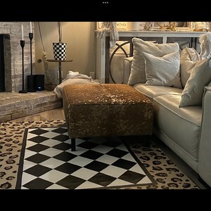 May include: A gold and brown cowhide ottoman with black legs sits on a black and white checkered rug with a leopard print border. The ottoman is in front of a white leather sofa with pillows.