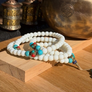Puede incluir: Cuentas de oración blancas con detalles turquesa y rojos sobre una superficie de madera. Un cuenco tibetano de bronce y dos ruedas de oración se encuentran en el fondo. Las cuentas tienen una borla.
