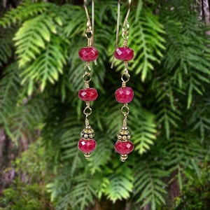 May include: A pair of dangle earrings featuring three faceted, deep pink ruby-colored beads suspended on gold-toned wire. Each earring includes a decorative gold-toned metal bead cap above the bottom stone and a simple gold-toned hook ear wire.
