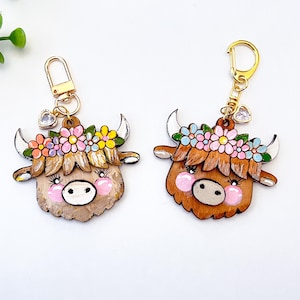May include: Two wooden Highland cow keychains. Each keychain features a brown cow face with a floral crown in pink, yellow, and blue. The cows have pink cheeks, white horns, and gold-colored hardware. A small heart charm hangs from the top.