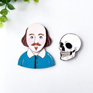 May include: Wooden cutouts of William Shakespeare and a skull. Shakespeare is depicted with a bald head, brown hair, a mustache, and a blue shirt. The skull is white with black eye sockets and teeth. The cutouts are likely for crafting or decoration.