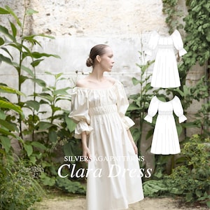 May include: A white off-the-shoulder dress with puffed sleeves and a gathered waist. The dress is knee-length and has a relaxed fit. The model is wearing the dress in a garden setting. The text "SILVERSAGA PATTERNS Clara Dress" is displayed below the model.