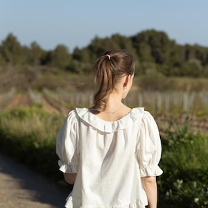 Pode incluir: Uma blusa branca com gola com babados e mangas curtas bufantes. A blusa tem bainha com babados e &eacute; combinada com cal&ccedil;as pretas. O fundo apresenta um caminho de terra e um campo com &aacute;rvores.