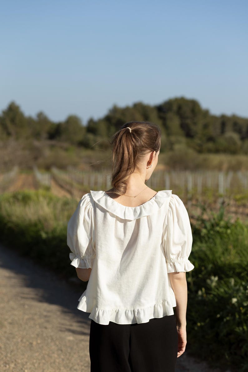 Pode incluir: Uma blusa branca com gola com babados e mangas curtas bufantes. A blusa tem bainha com babados e &eacute; combinada com cal&ccedil;as pretas. O fundo apresenta um caminho de terra e um campo com &aacute;rvores.