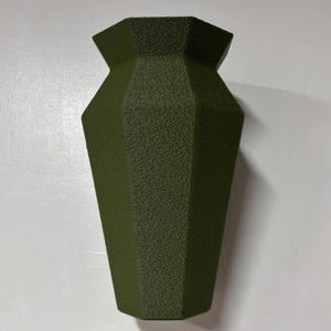May include: A dark green, geometric vase with a textured surface. The vase is tall and narrow, with a rounded base and a slightly flared opening.