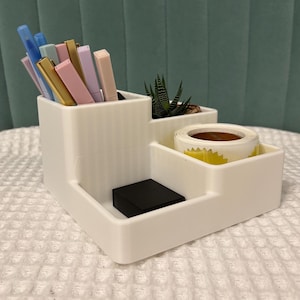 May include: White desktop organiser with four compartments. The organiser has a compartment for pens and pencils, a compartment for a small plant, a compartment for tape, and a compartment for small items.