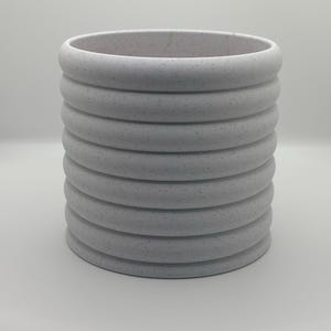 Puede incluir: Un macetero de cerámica blanca con un diseño texturizado y acanalado. El macetero es cilíndrico y tiene un borde liso y redondeado.