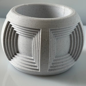Puede incluir: Una maceta esférica gris claro con una superficie texturizada. La maceta presenta un diseño geométrico de cuadrados y rectángulos empotrados alrededor de su circunferencia. La abertura superior es circular.