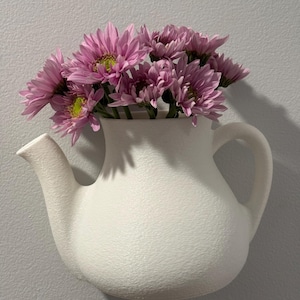 Peut inclure: Une théière en céramique blanche à surface texturée, utilisée comme vase pour un bouquet de fleurs roses.