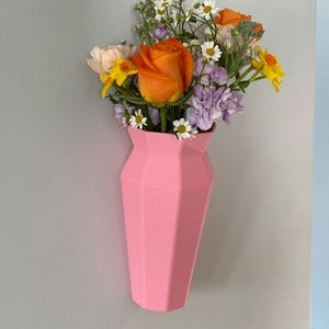 May include: A pink geometric vase with a bouquet of flowers, including orange roses, white daisies, and purple flowers.