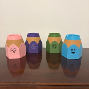 Könnte beinhalten: Vier buntstiftförmige Behälter mit verschiedenen Emoji-Gesichtern. Die Behälter sind rosa, lila, grün und blau, mit einem braunen Stiftkörper. Jedes Gefäß hat ein anderes Emoji-Gesicht: schlafend, traurig, lächelnd und sternenäugig.